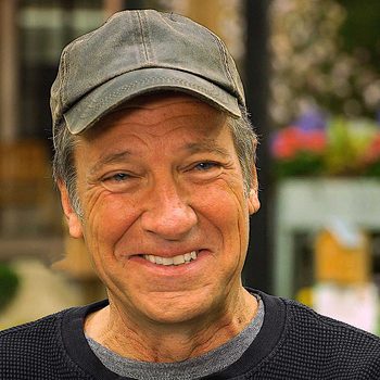 A smiling man wears a cap, showcasing weathered features. He stands outdoors with a blurred background of greenery and colorful flowers.