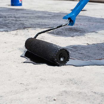 A paint roller applies black material on a light concrete surface, with puddles forming around it, suggesting a fresh coating process in progress.