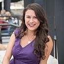 A smiling woman with long, wavy hair sits casually, wearing a purple dress, against a blurred background of people and a building.