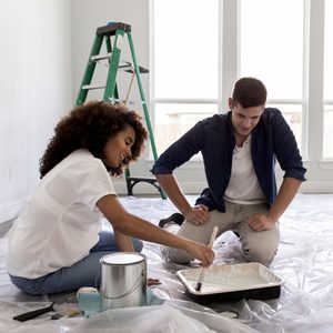 A woman paints a tray with a brush while a man observes, both sitting on a covered floor in a brightly lit room with a ladder nearby.