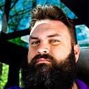 A man with a thick beard gazes confidently at the camera, seated in a vehicle with sunlight filtering through trees in the background.