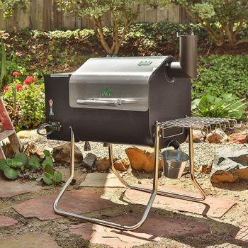 A black smoker grill sits on metal legs, surrounded by a landscaped garden with flowers and greenery, ready for outdoor cooking.