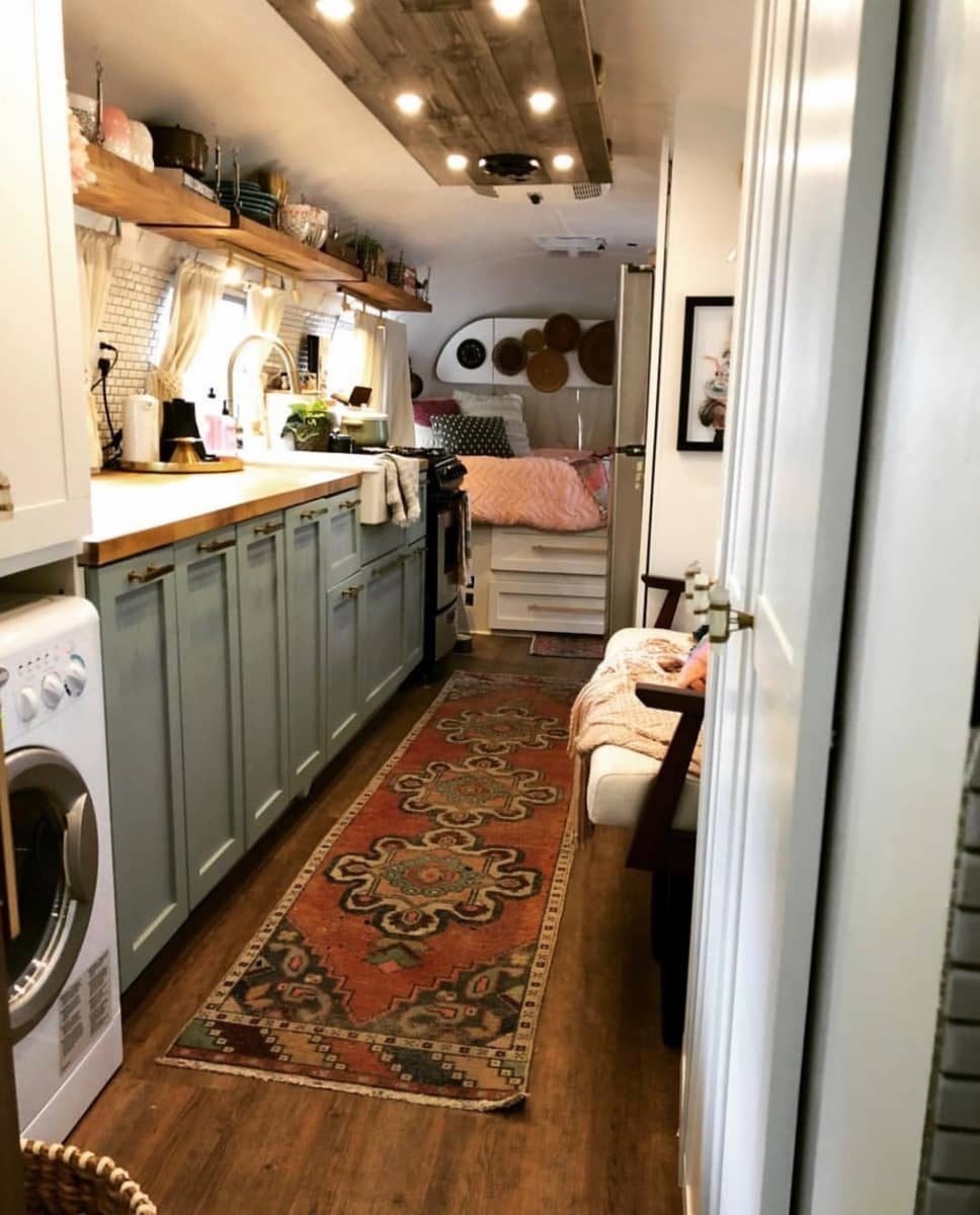 A cozy, narrow interior features a kitchen area with appliances, a colorful rug running throughout, and a bed visible in the background.
