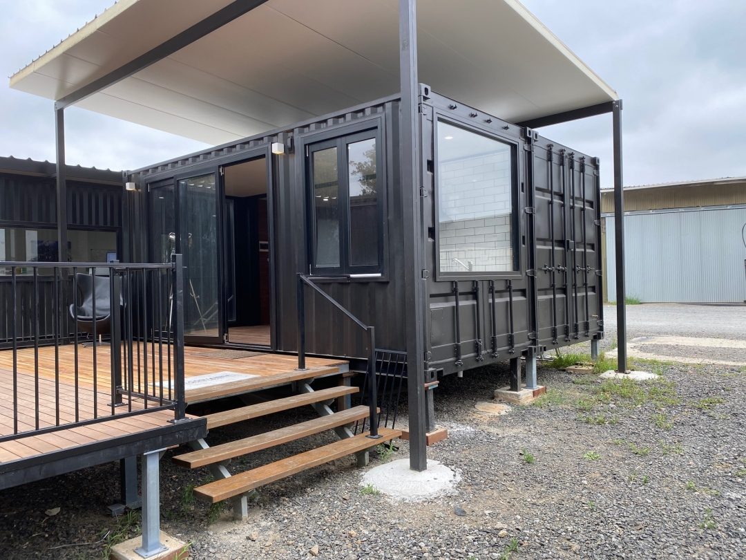 A black shipping container house showcases large windows and a deck, with steps leading up to the entrance, situated in a gravel area under overcast skies.
