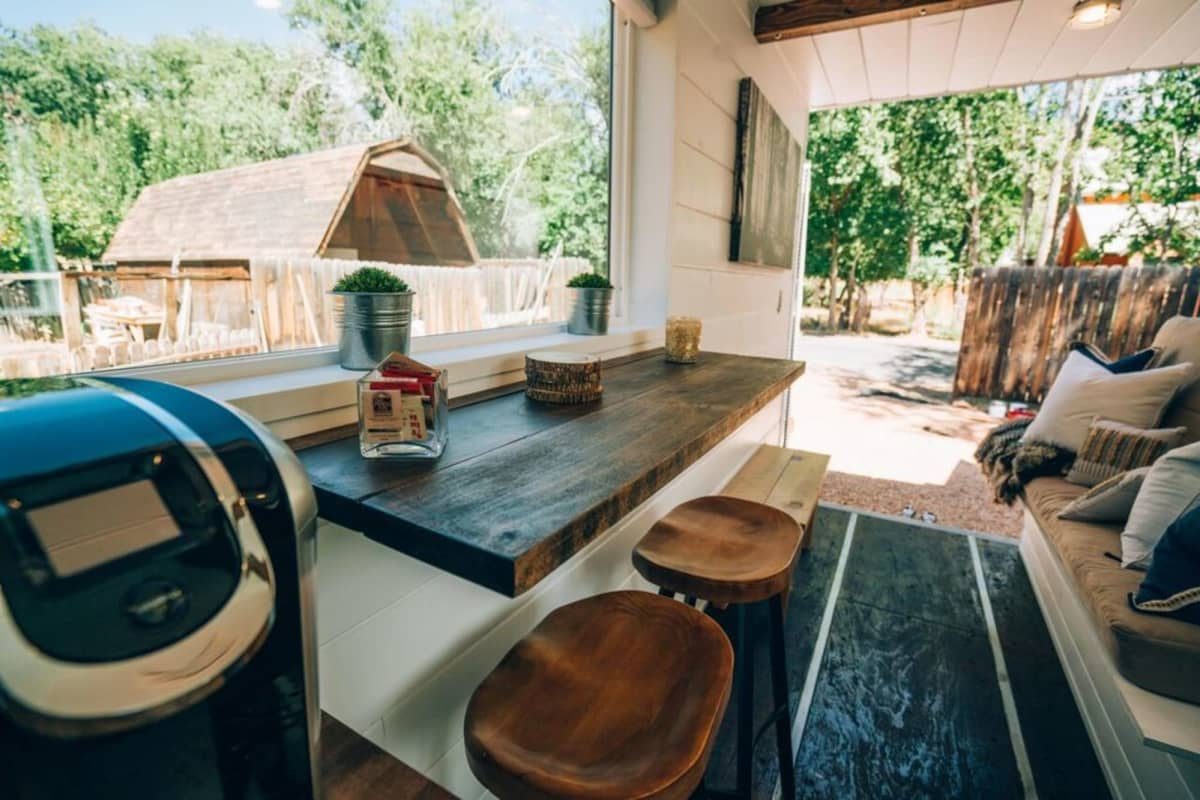 A wooden countertop with two stools sits inside a bright space, featuring a coffee maker, decorative plants, and large windows revealing an outdoor view.