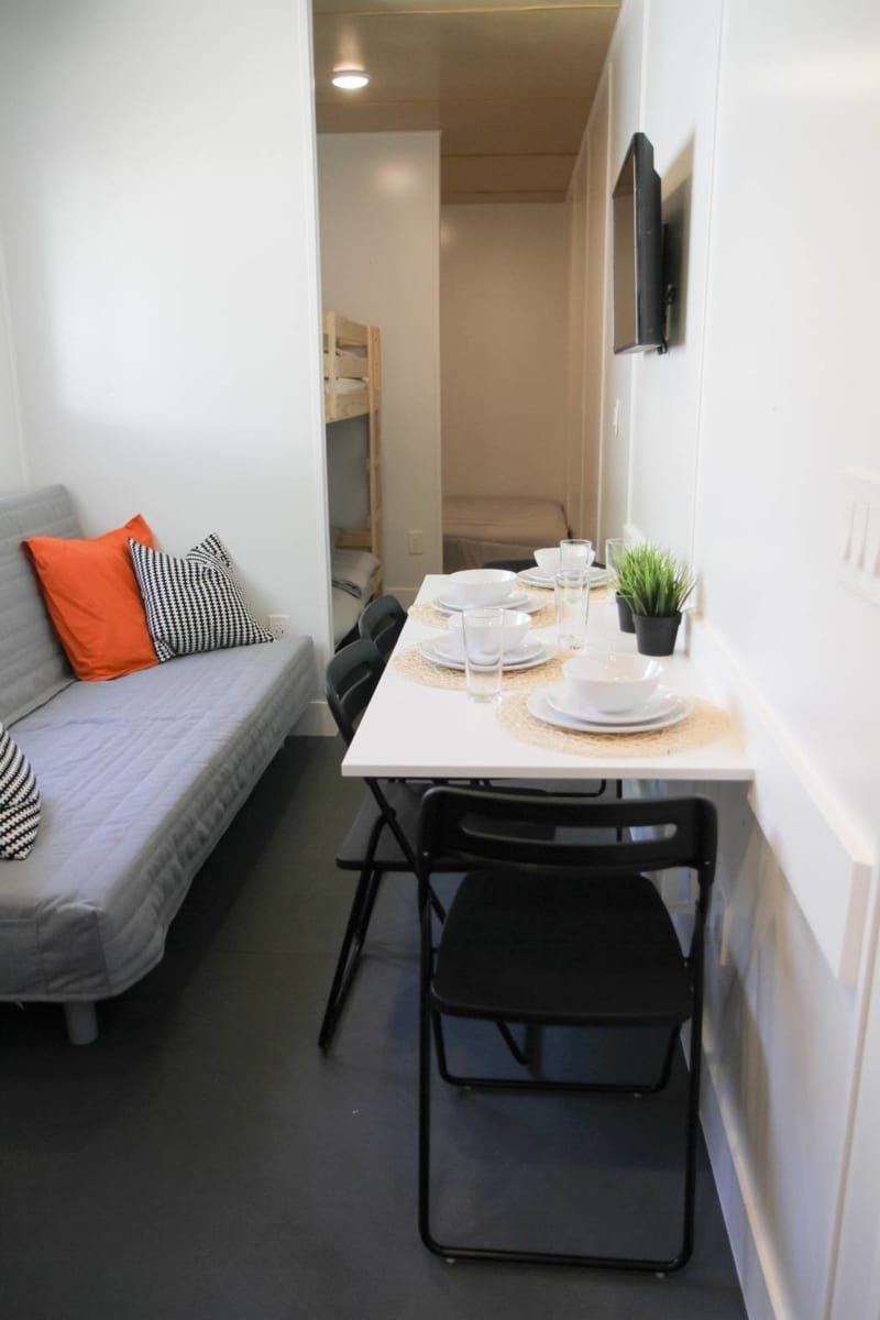 A gray sofa, decorated with pillows, sits beside a dining table set with plates and glasses, in a small, modern, well-lit living space.