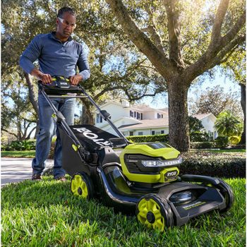A person is pushing a green and black lawn mower across a grassy yard near a house, surrounded by trees and sunlight.