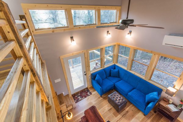 A blue couch occupies a cozy wooden living room with large windows, illuminating a space that connects to an outdoor deck in a tranquil forest setting.