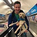 A person holds a puppy while standing on an escalator in a subway station, with advertisements visible in the background.