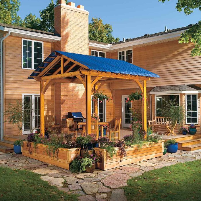 A wooden gazebo with a blue roof stands in a garden, surrounded by potted plants, stone pathways, and patio furniture near a house with beige siding.