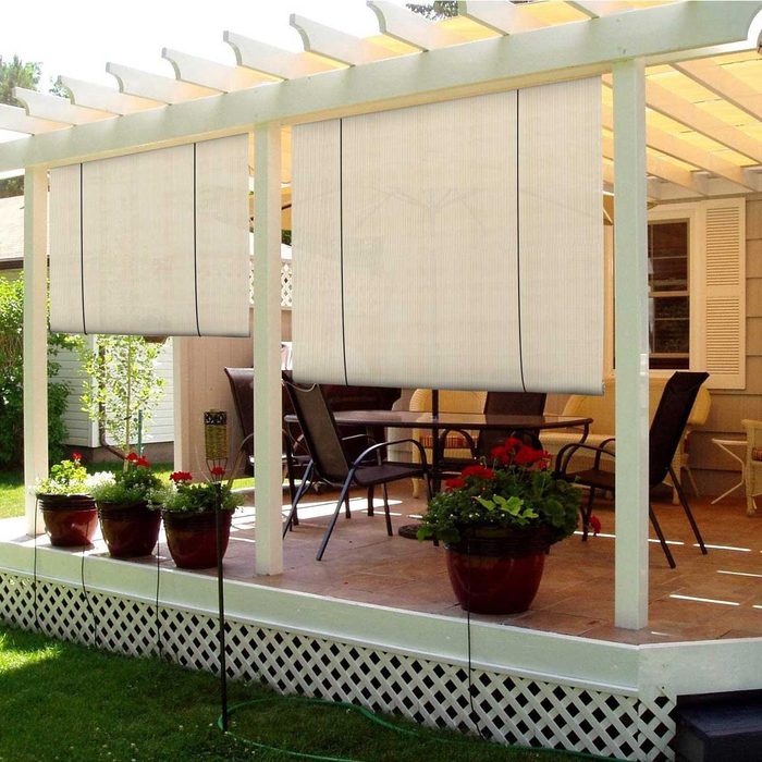 Vertical shades hang down in a covered patio area, shielding a seating arrangement with plants below, surrounded by greenery and a cozy atmosphere.