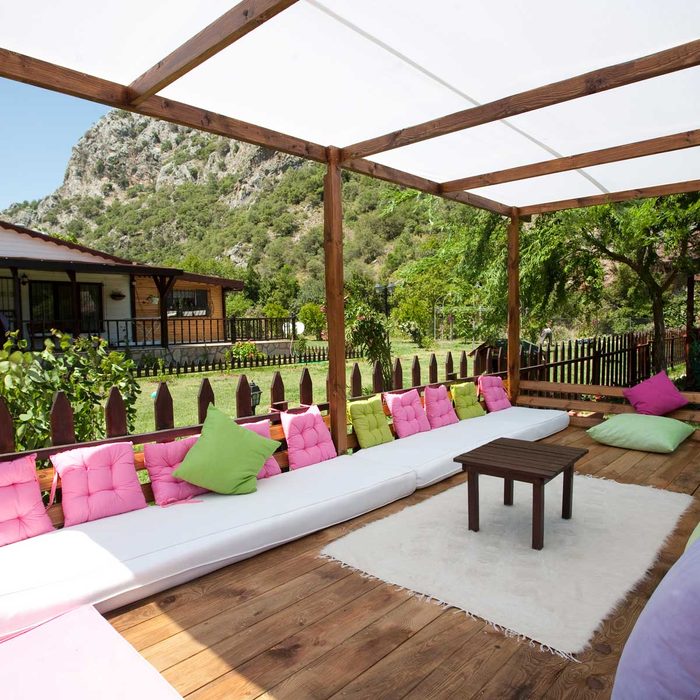 A wooden pergola houses a white couch with colorful cushions. A small table sits on a rug, surrounded by a green lawn and mountains.