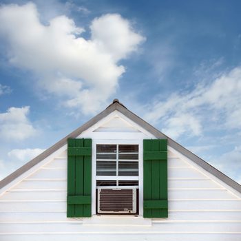 White house corner features an air conditioner in a window, flanked by green shutters, against a blue sky with clouds.