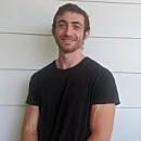 A young man stands smiling, wearing a black t-shirt. He has short hair and is positioned in front of a white wall.