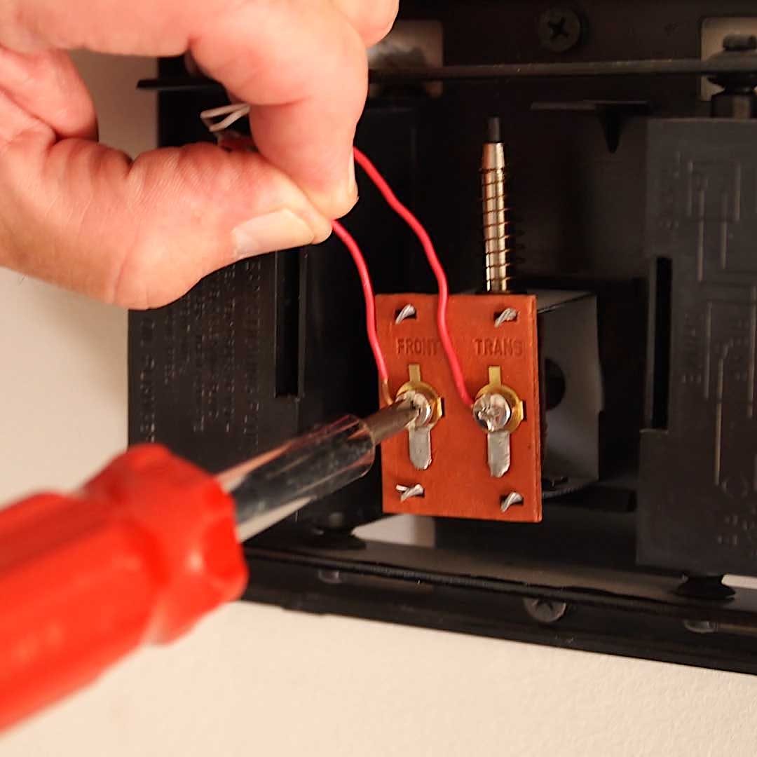 A hand holds red wires while using a screwdriver to connect them to a terminal block mounted on a wall. The background is neutral.