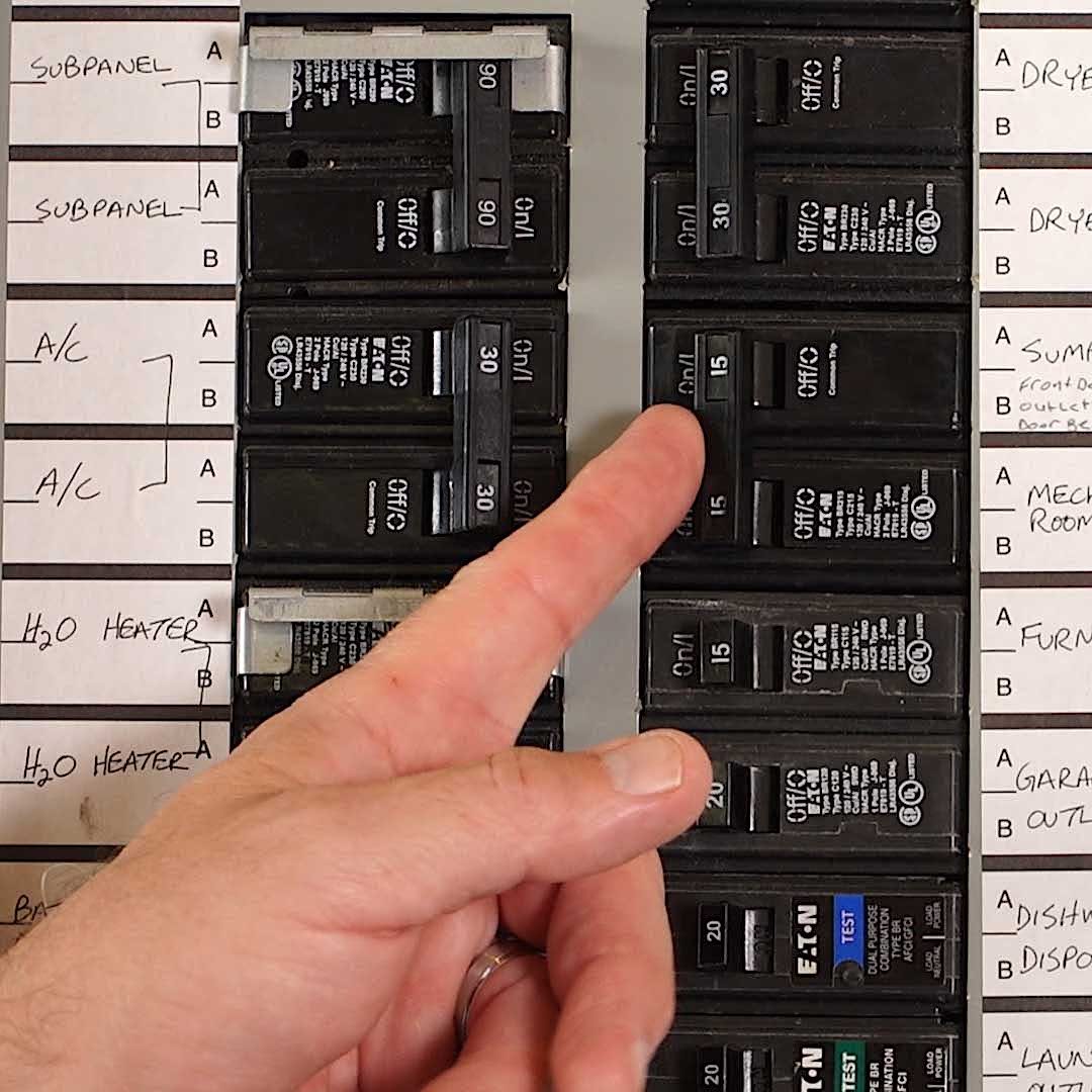 A hand points to circuit breakers on a panel, which are labeled for different electrical appliances in a home setting.