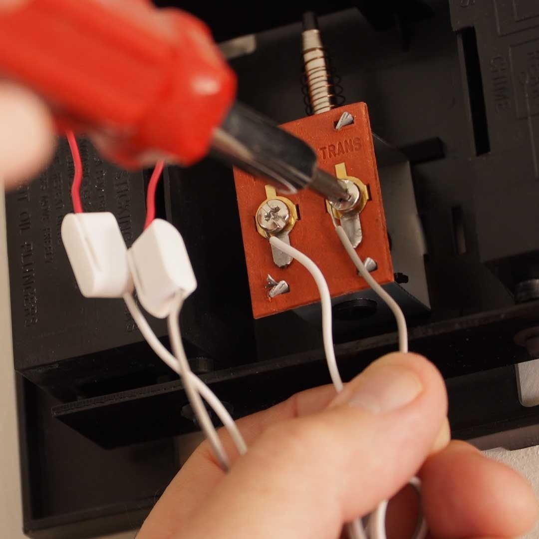 A hand holds a screwdriver, connecting wires to a circuit board with terminals, set against a dark background, likely part of electrical installation or repair.