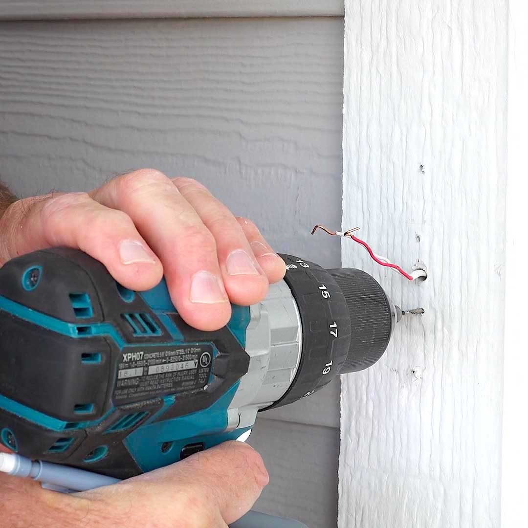 A person uses a cordless drill to create a hole in a white wall, with exposed electrical wires nearby.