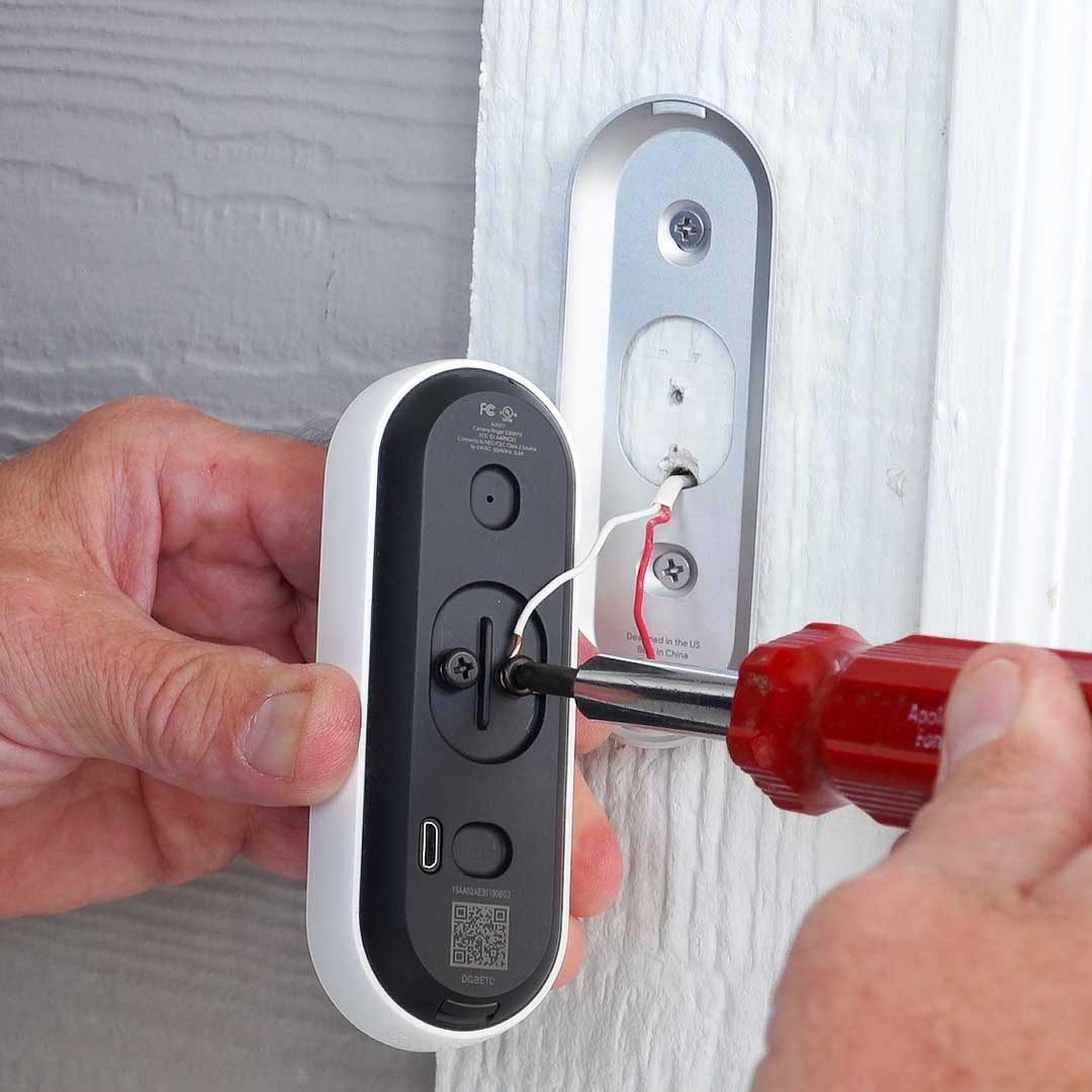 A hand holds a screwdriver, installing a black-and-white doorbell onto a wall, with wires connected. The environment appears to be a home entryway.