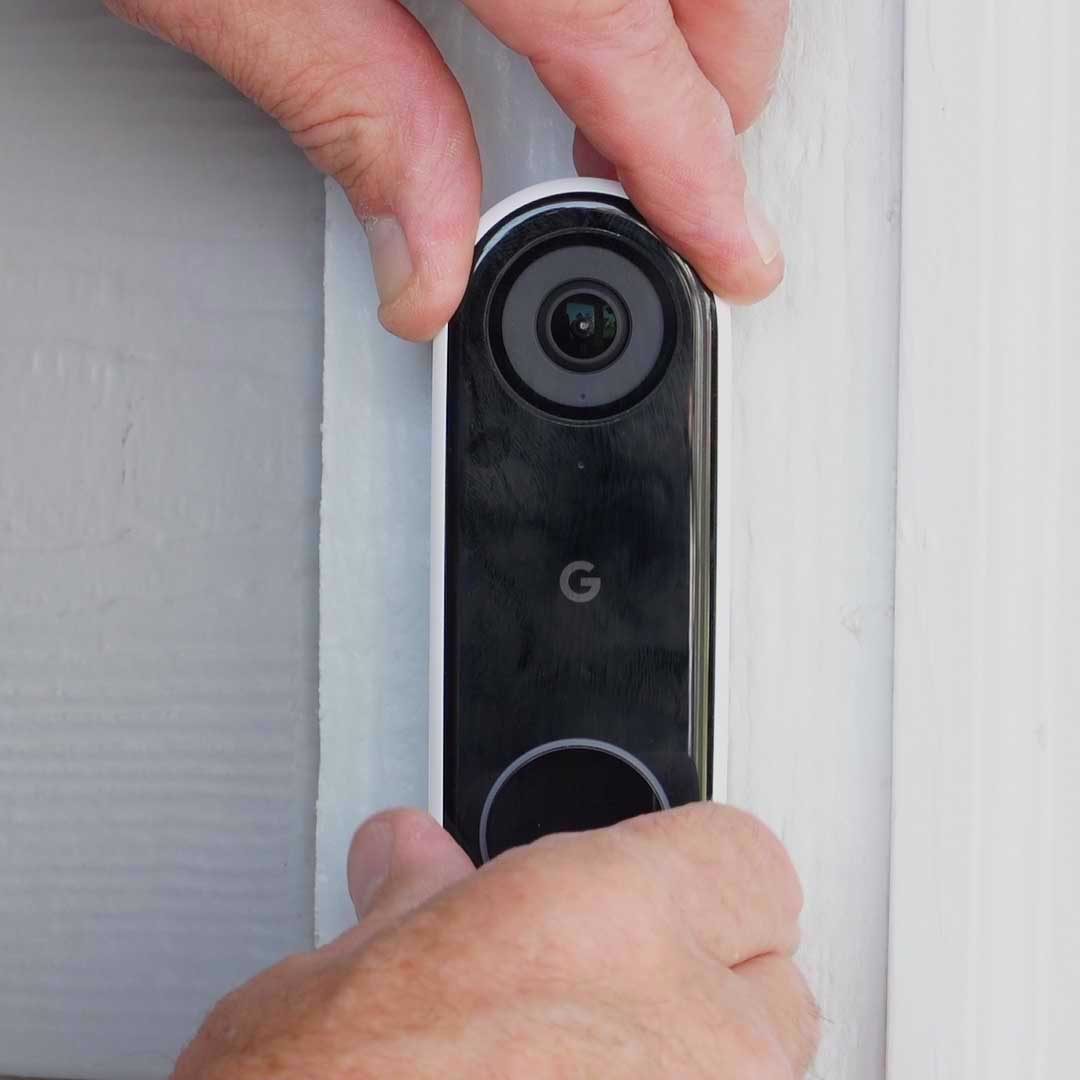 A person is installing a black smart doorbell on a light-colored wall, positioning it carefully with hands gripping the device.