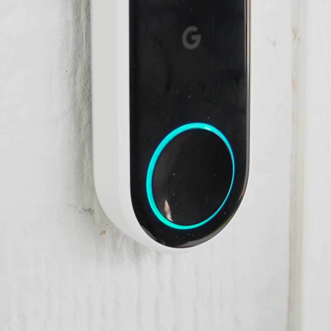 A doorbell with a circular, illuminated button illuminates as it's pressed, mounted on a textured white wall in a home setting.