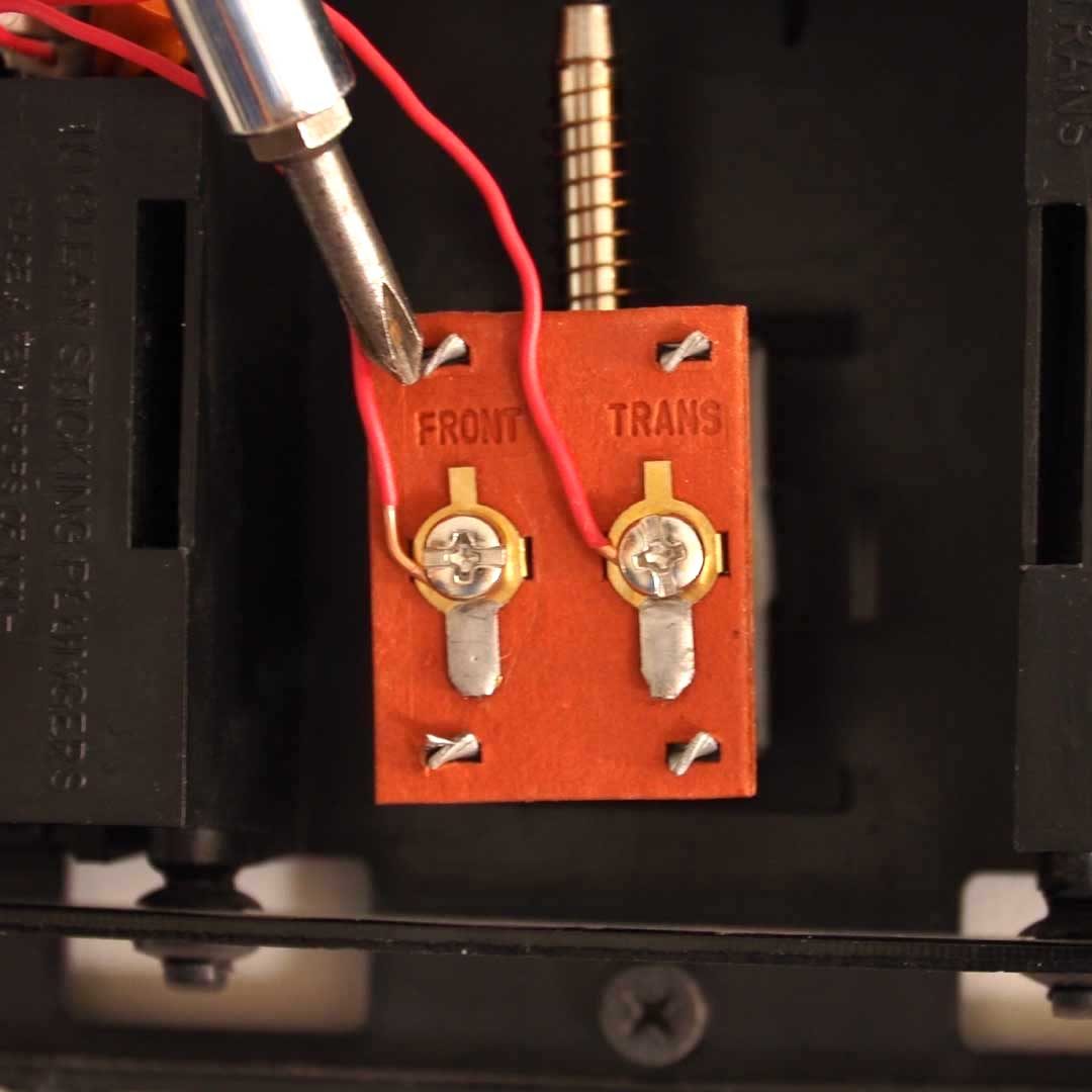 A screwdriver is positioned over a circuit board with two gold terminals labeled "FRONT" and "TRANS," connected by red wires in an electronic device.