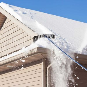 A snow shovel clears snow from a roof's edge, causing snow to cascade down, against a backdrop of a clear blue sky and a house's siding.