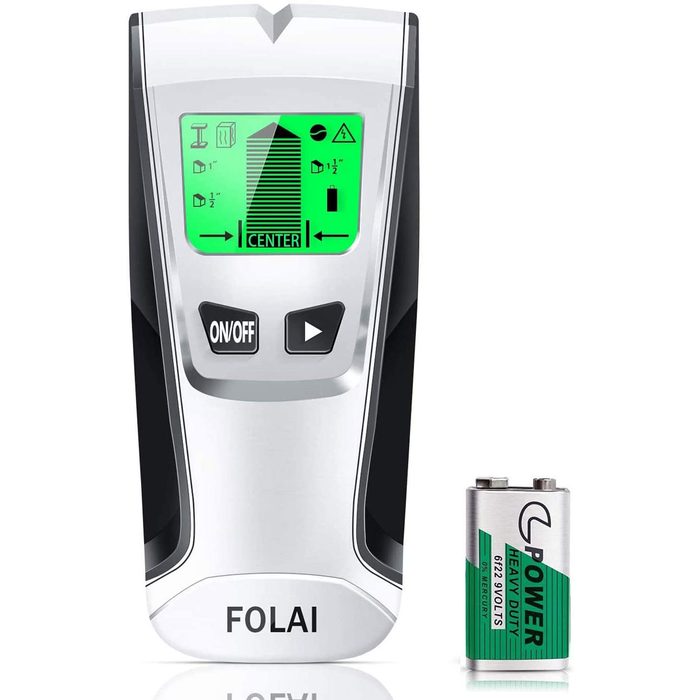 A digital stud finder displays a green screen with measurement indicators while resting beside a battery, suggesting it