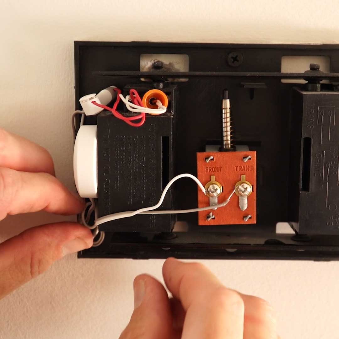A hand connects wires to a red terminal block mounted on a black wall plate, with additional wires and components visible in the background.