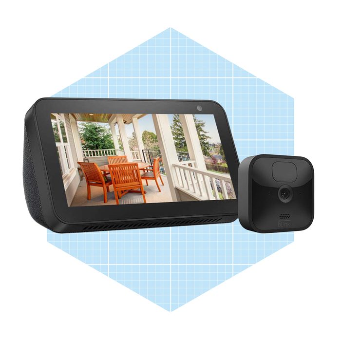 A smart display and a camera are positioned together, showcasing a view of a residential patio with wooden furniture under a covered area, surrounded by greenery.