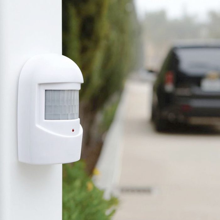 A white motion detector is mounted on a wall, monitoring movement near a driveway lined with greenery, alongside a parked black vehicle in the background.