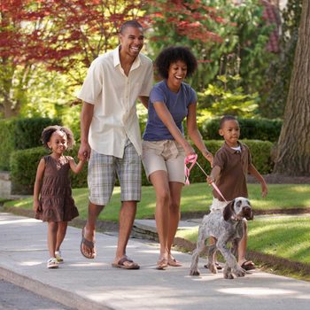 A family walks together on a path, smiling and holding hands, while a child walks a dog, surrounded by greenery and trees.