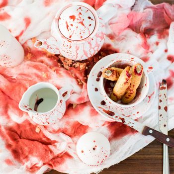 A blood-stained tea set is spilled on a red-patterned cloth, with a plate holding food resembling fingers, contrasting with the wooden table underneath.