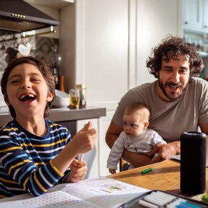 A smiling boy holds a marker while sitting at a table with a baby and a man, in a kitchen filled with natural light.