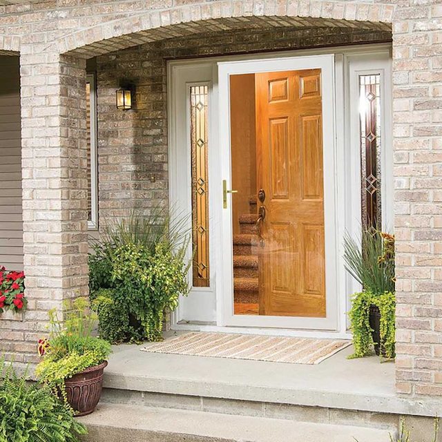 A white front door with decorative side panels opens inward, revealing a stairway inside. Potted plants frame the entrance on a stone porch.