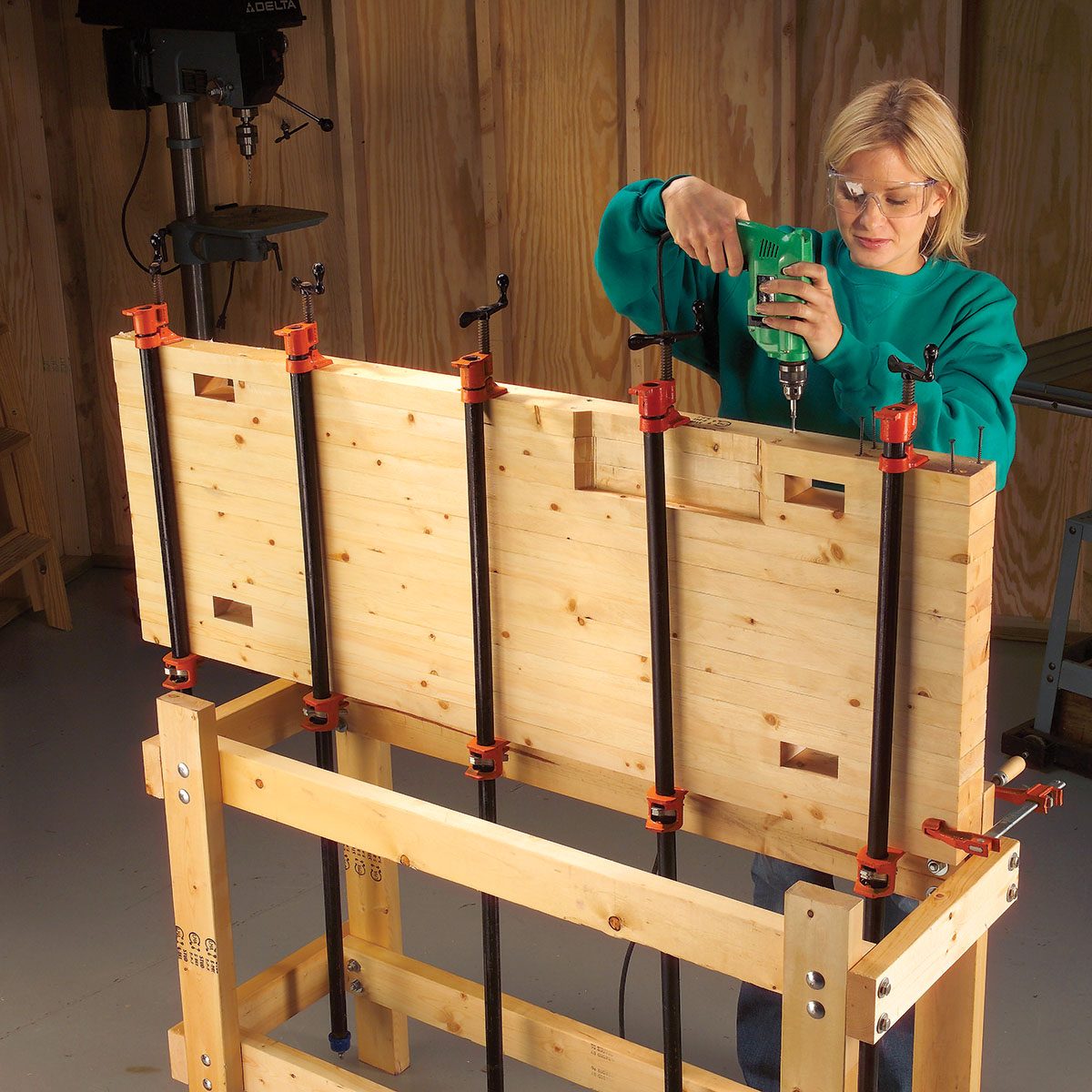 A woman drills into a clamped wooden board on a workbench in a workshop with wooden walls and tools in the background.