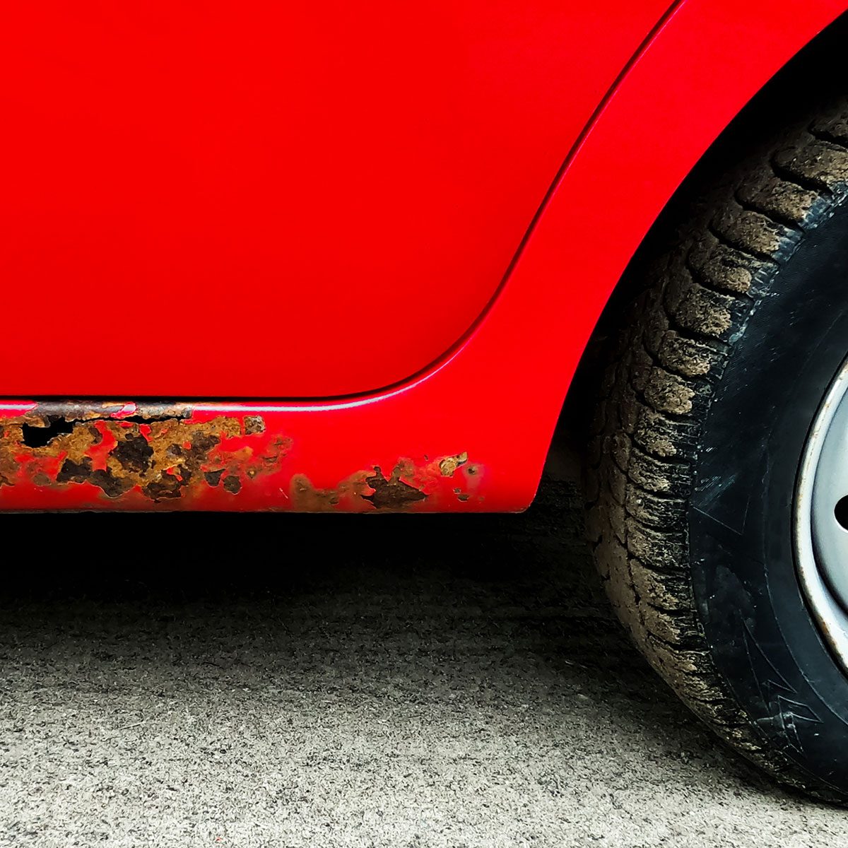 A red car with rust spots along the lower body is parked on a rough, gray surface, and its tire shows signs of wear and mud.