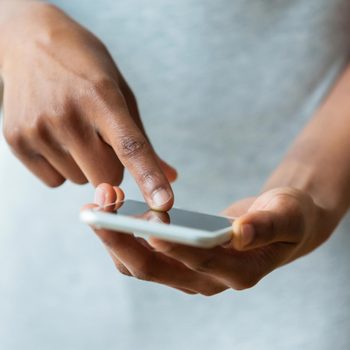 A hand holds a smartphone, with a finger touching the screen. The background is softly blurred, suggesting an indoor setting.