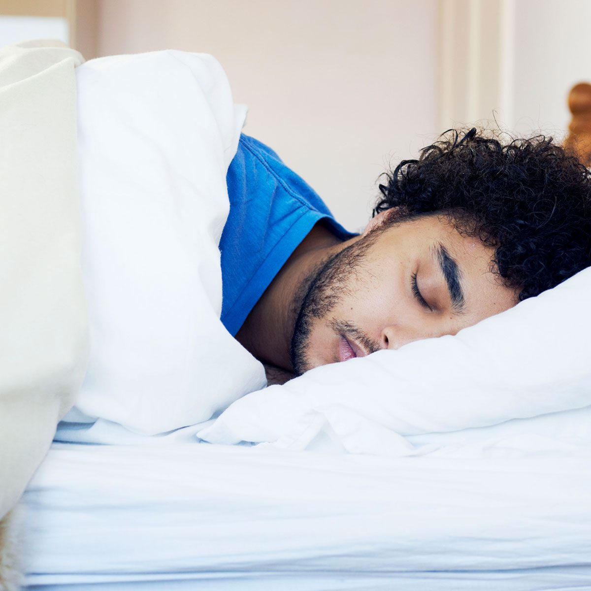 A young man sleeps peacefully on a bed, nestled among white sheets and pillows, in a softly lit indoor space.