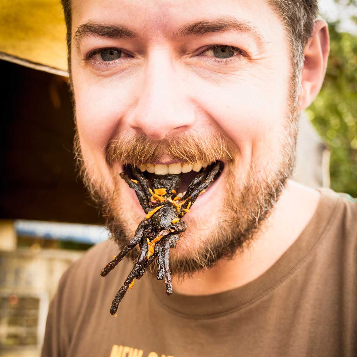 A man smiles while having a dark, spider-like food item protruding from his mouth, set against a blurred outdoor background.