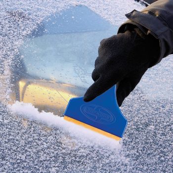A blue ice scraper is being used to remove snow from a car windshield, surrounded by a snowy environment, indicating cold winter conditions.