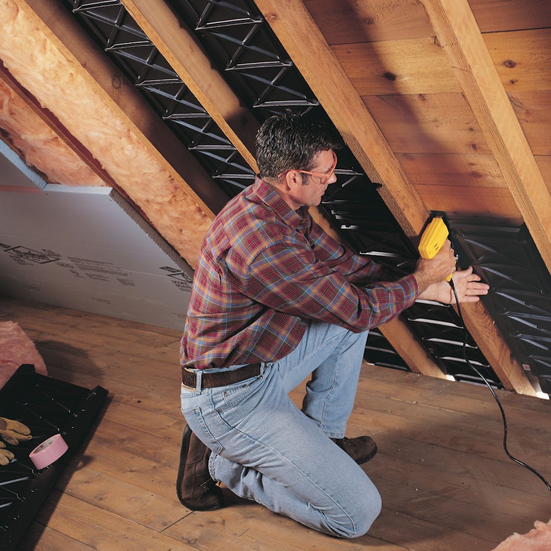 A man kneels while using a power tool to attach materials in an attic with wooden beams and insulation visible in the surroundings.