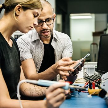 Two people, a man and a woman, are focused on a device being repaired at a cluttered workspace, sharing technical insights while surrounded by tools and equipment.