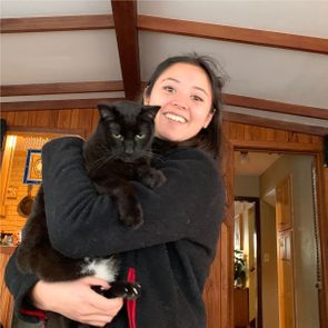 A woman smiles while cradling a black cat in her arms, standing indoors with wooden beams and an open doorway in the background.
