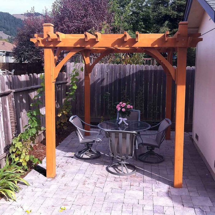 A wooden pergola shades a circular table with four chairs, adorned with a pink flower bouquet, nestled in a garden surrounded by a wooden fence.