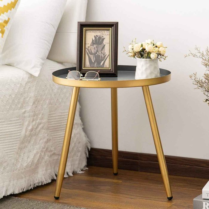 A round table with gold legs supports a framed picture, glasses, and a small vase of flowers, positioned beside a bed, against a neutral wall.