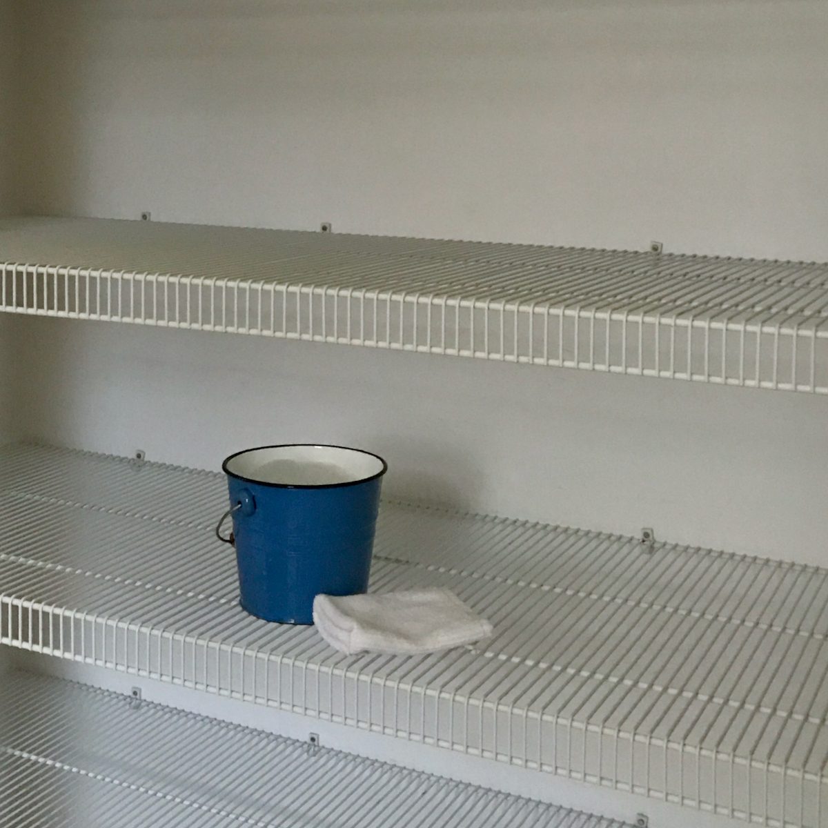A blue bucket sits on a white wire shelf, beside a folded white cloth, in an empty storage area with multiple shelves.