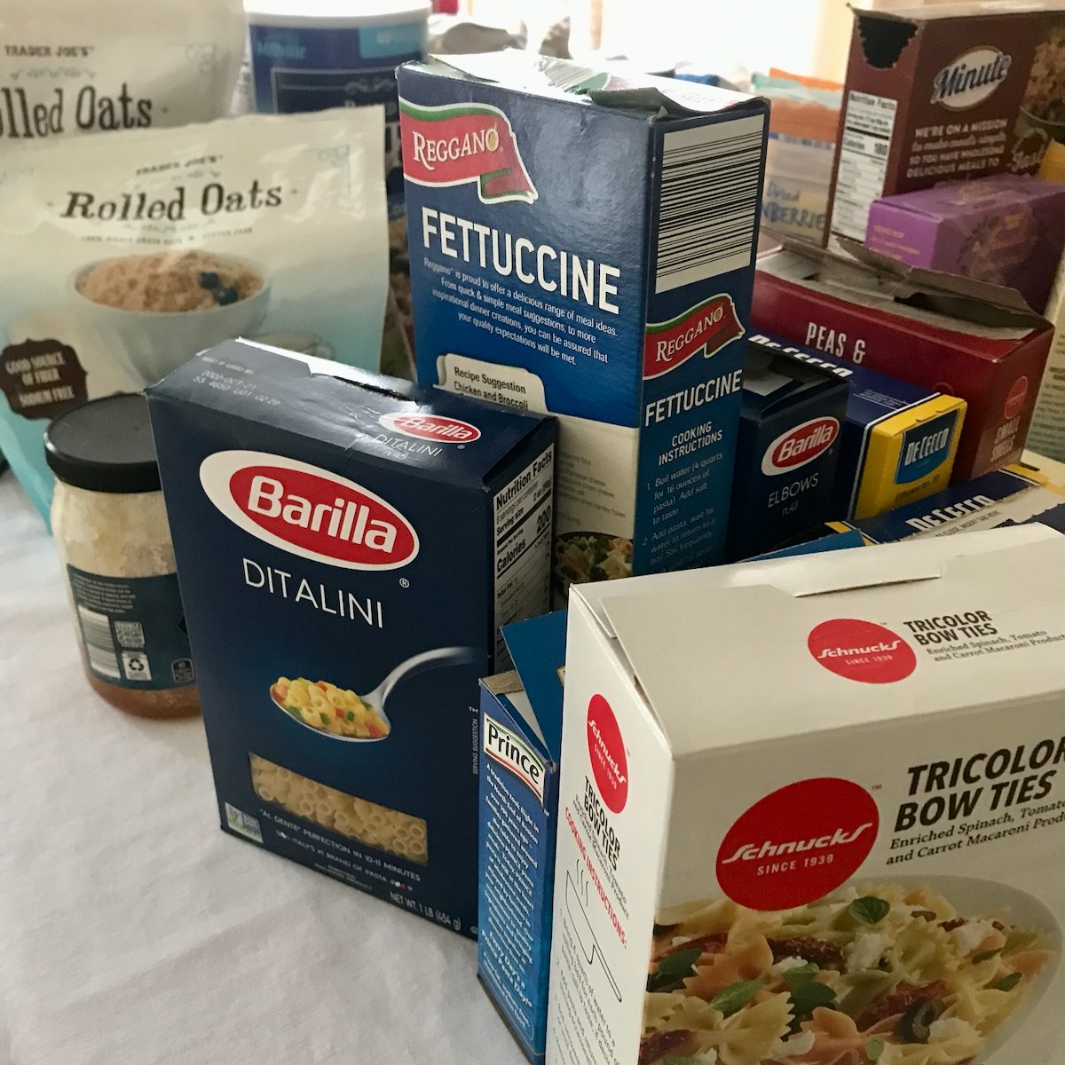 Boxes of various pasta types are stacked together on a table, with labels visible, amidst containers of oats and additional food items in the background.