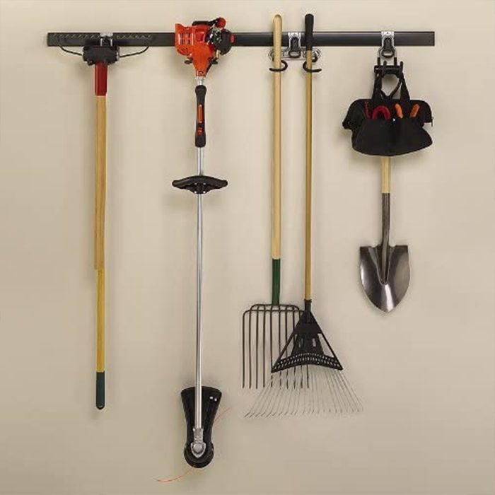Gardening tools hang on a wall-mounted rack, including a trimmer, shovel, rake, and a tool bag, arranged for easy access in a garage or shed.