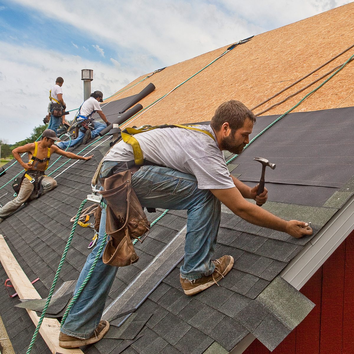 Workers install shingles on a sloped roof, using tools and safety harnesses, under a partly cloudy sky with a near structure in the background.
